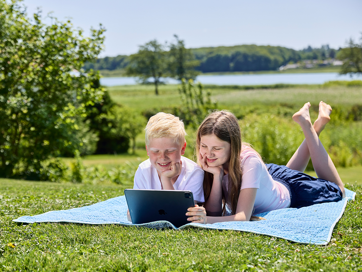 Bror og søster ligger på et tæppe i haven og ser på tablet