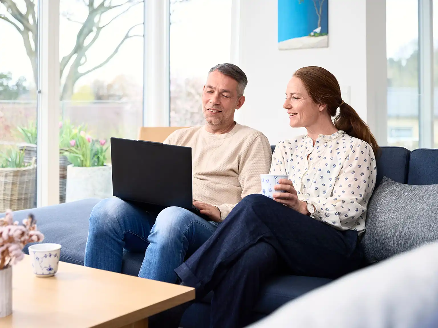 Mand og kvinde sidder sammen i sofaen manden holder en bærbar computer som de begge kigger fokuseret på kvinden holder en kop