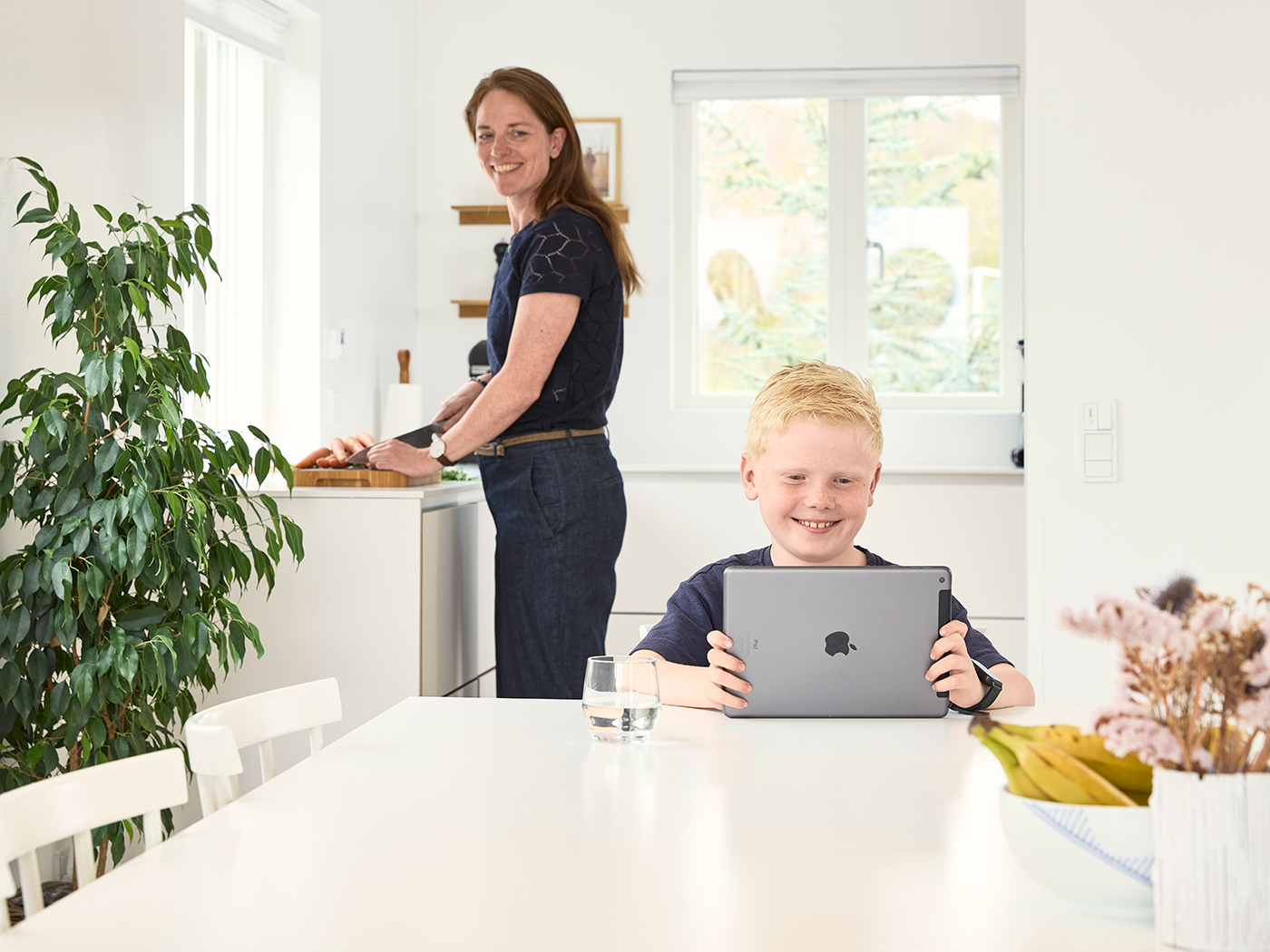 Søn sidder med tablet ved spisebordet mens hans mor kigger over til ham fra køkkenet
