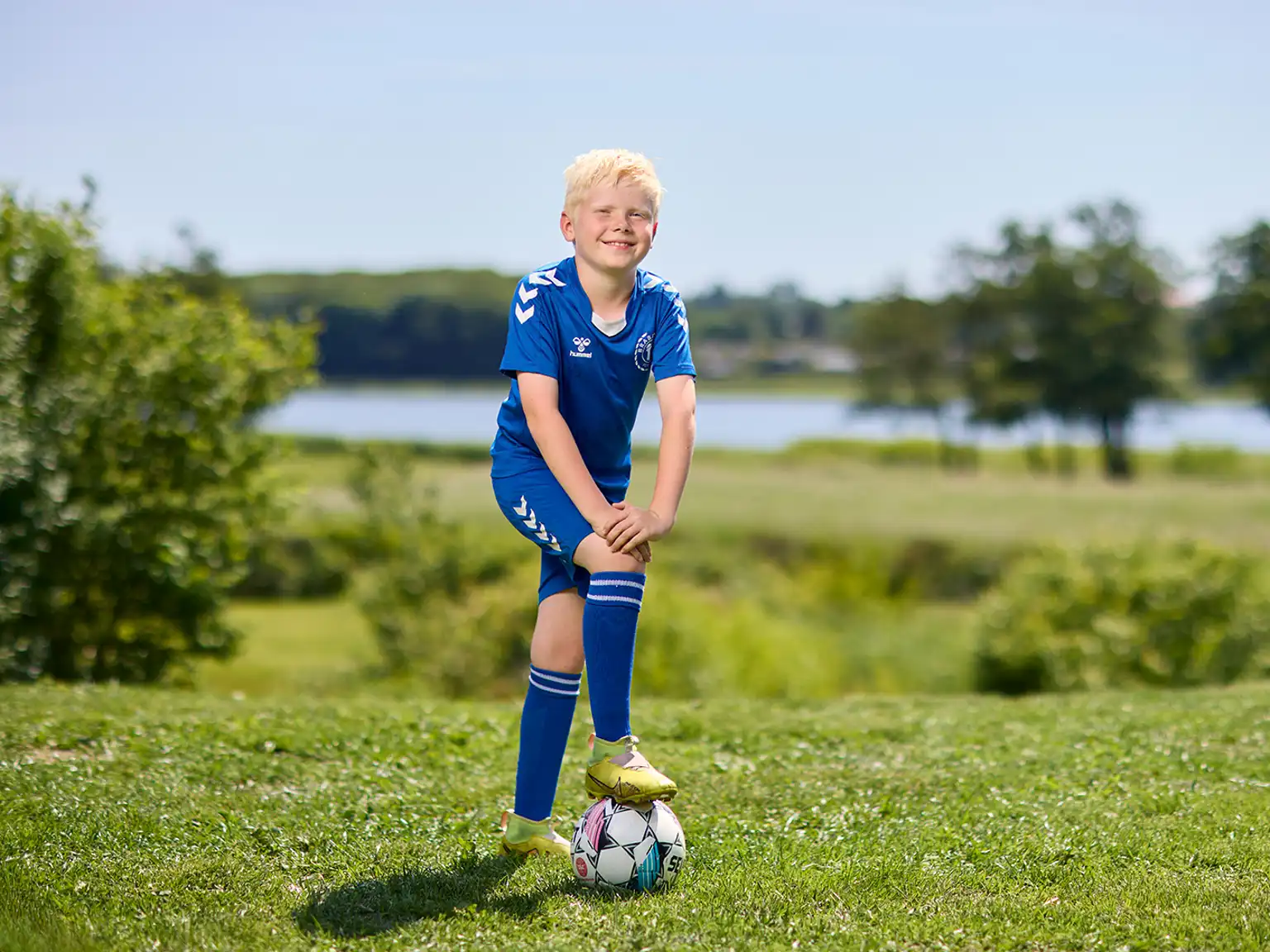 Dreng står i haven i fodboldtøj med foden på fodbold og smiler