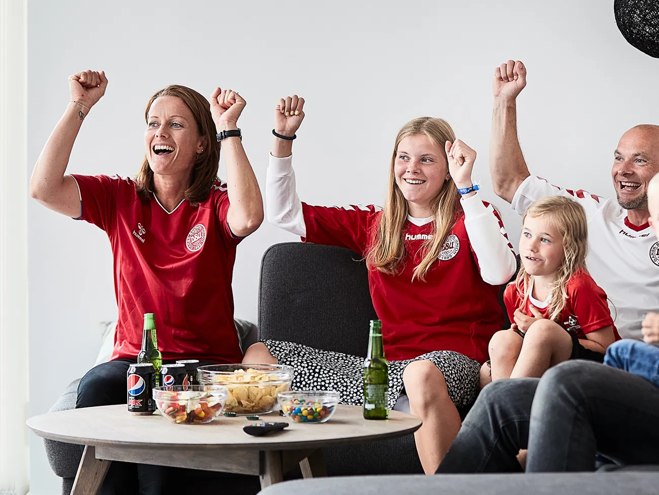 Familie sidder i sofaen og ser sport sammen, de jubler med armene over hovedet og har snacks på sofabordet