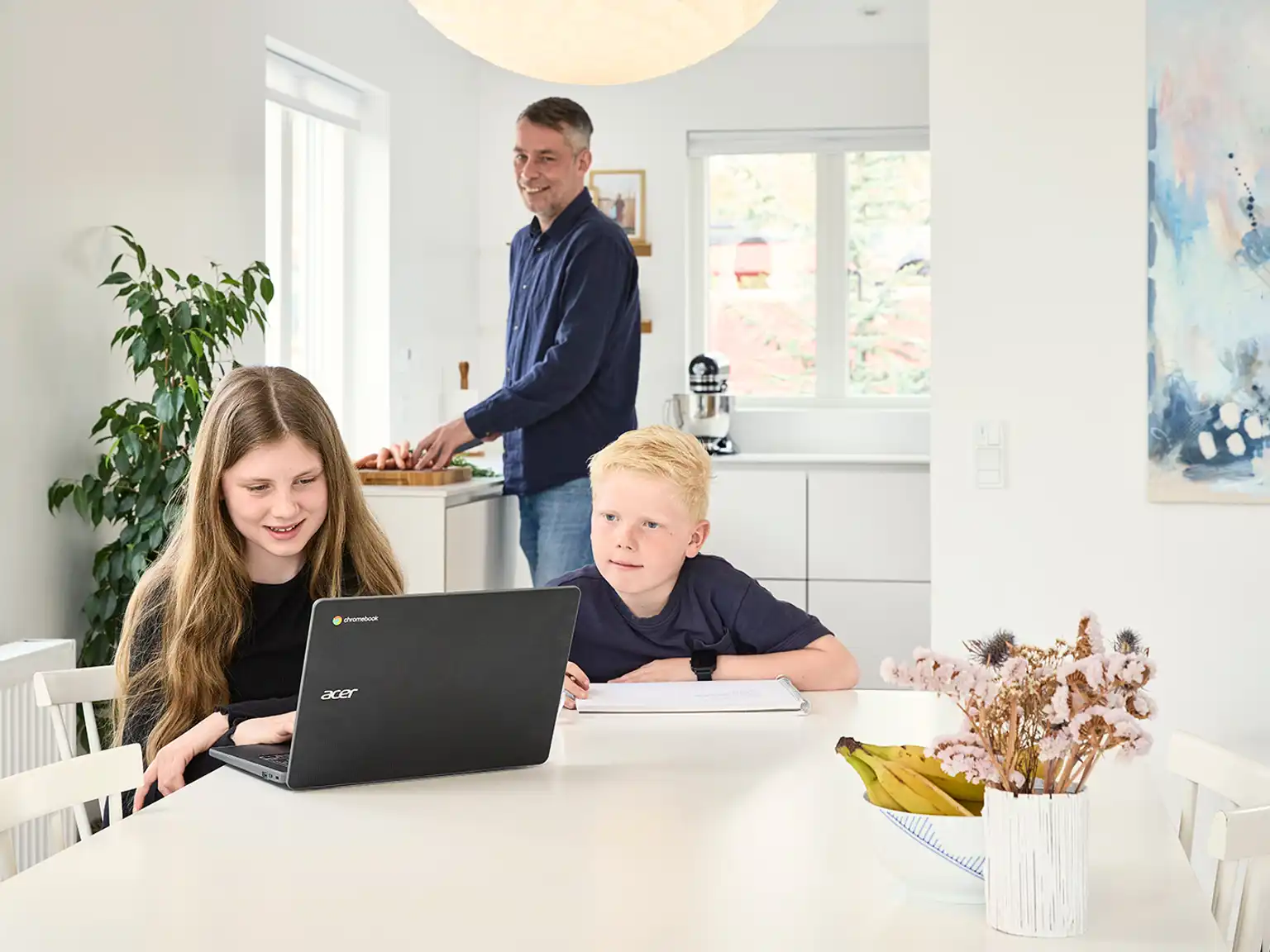 Pige og dreng sidder sammen og laver lektier mens deres far laver mad i køkkenet