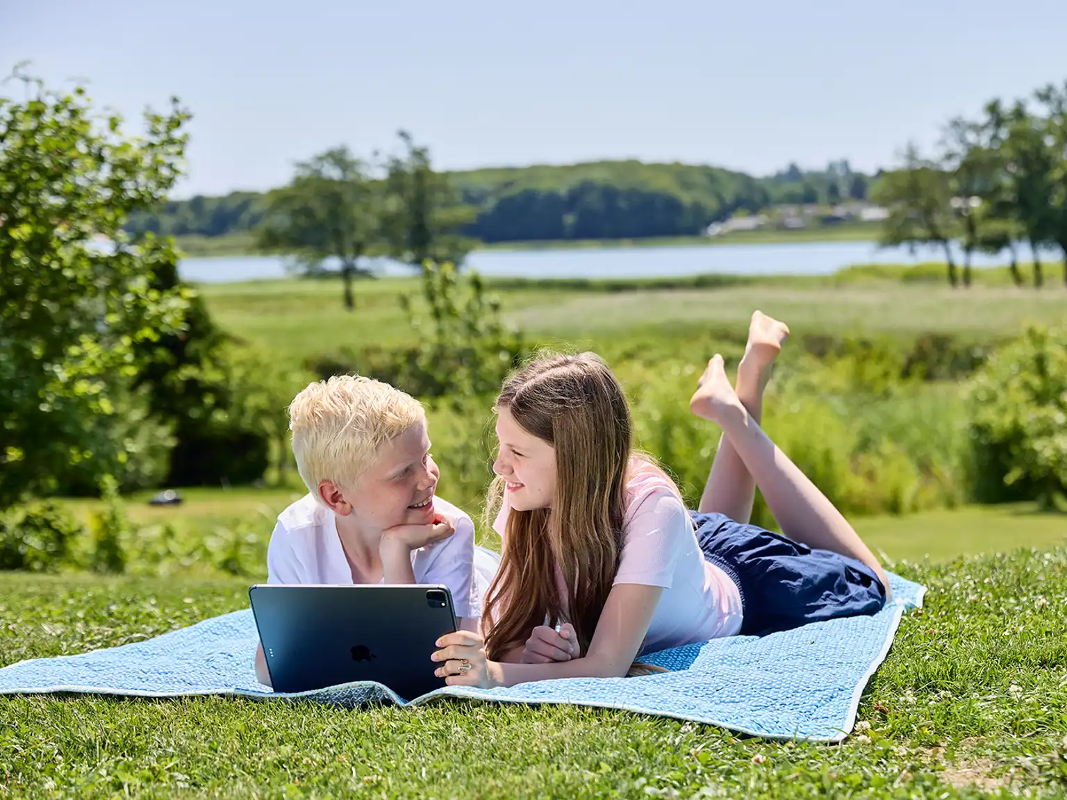 Dreng og pige ligger på et tæppe i haven med tablet i hånden