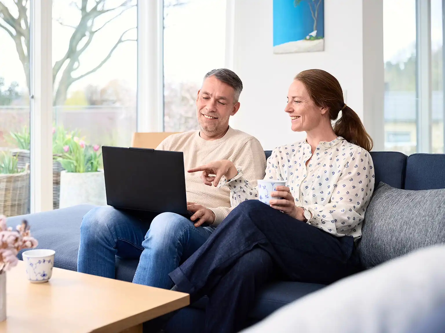 Mand og kvinde sidder sammen i sofaen og smiler manden holder en bærbar computer som de begge kigger på kvinden peger på computeren
