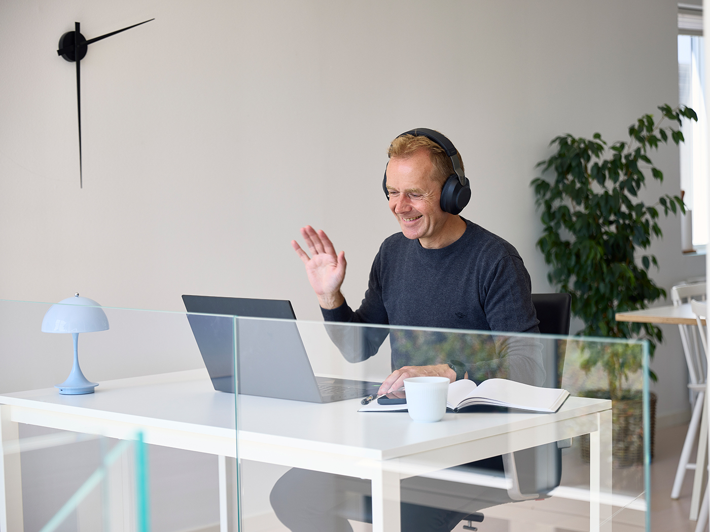 Mand sidder ved et skrivebord og vinker til kameraet på sin computer