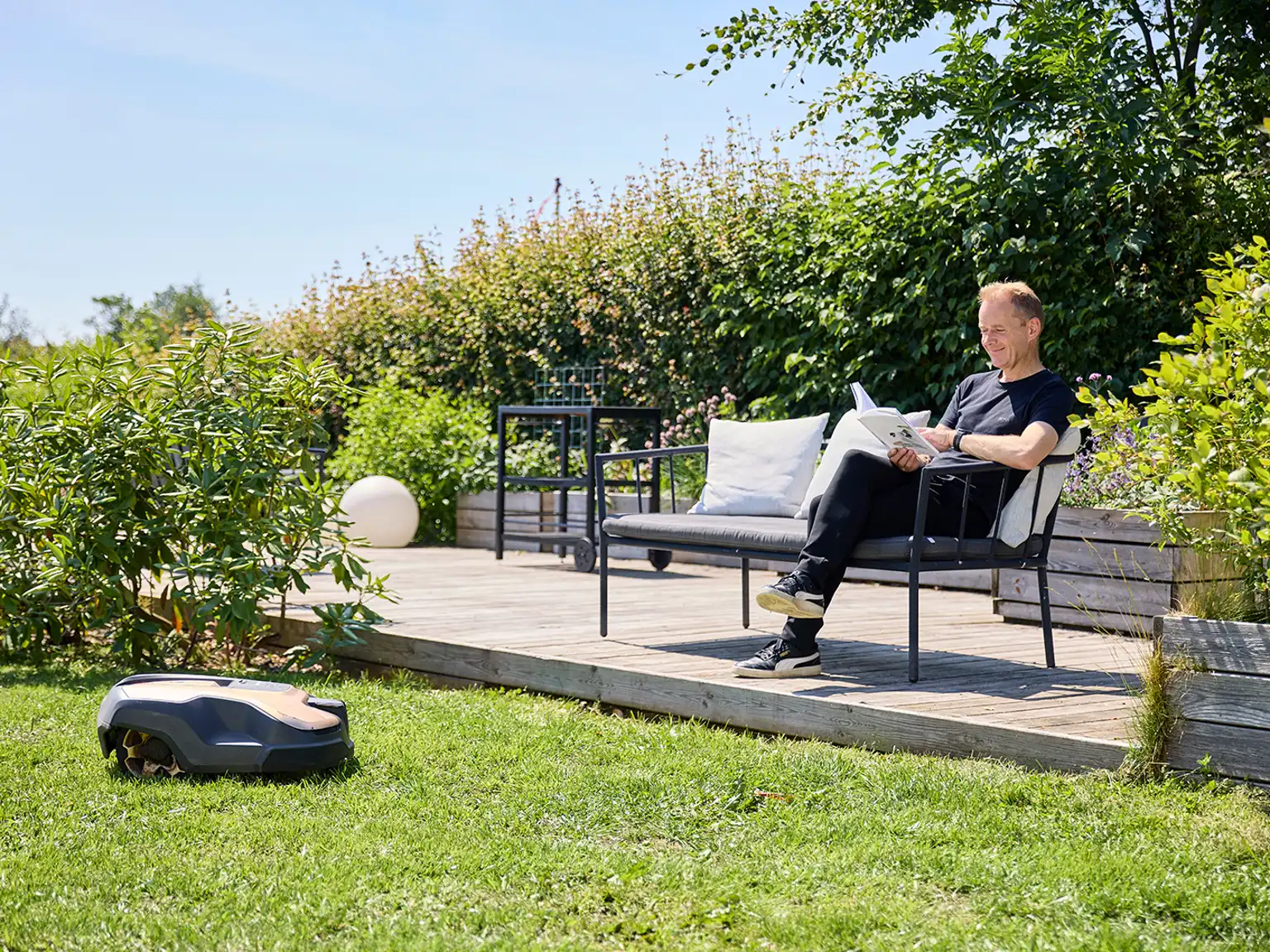 Mand sidder i en havestol og læser en bog i solen mens robotplæneklipperen klipper græs