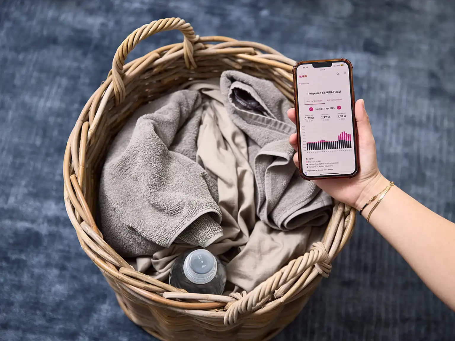 Mobil med overblik over elpriserne time for time holdes i en hånd ind over en vasketøjskurv fyldt med vasketøj og en flaske med vaskemiddel