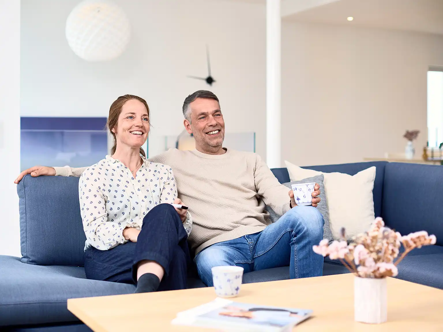 Kvinde og mand sidder tæt sammen i sofaen og griner kvinden holder en fjernbetjening og manden holder en kop