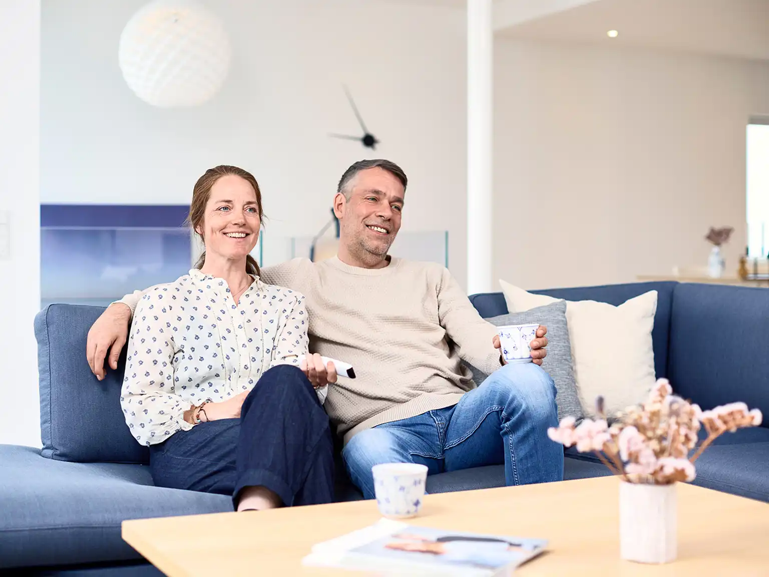 Kvinde og mand sidder afslappet sammen i sofaen manden har en arm rundt om kvinden hun holder fjernbetjeningen til tvet