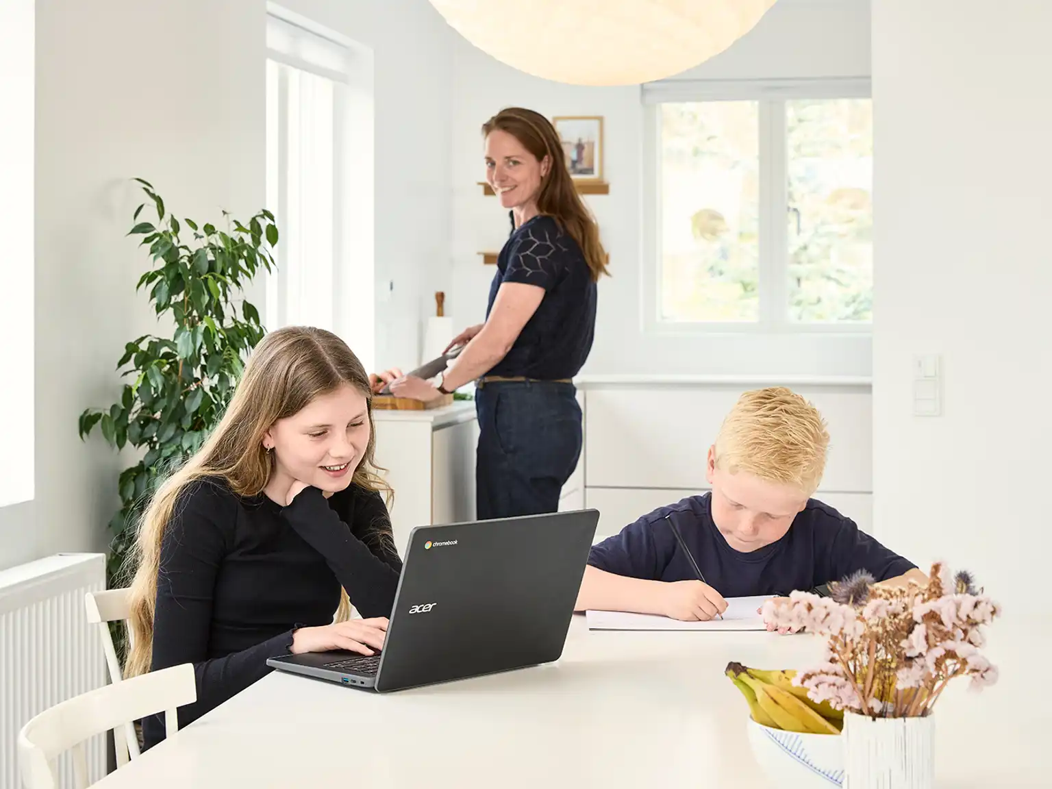 Pige og dreng sidder ved spisebordet og laver lektier mens deres mor kigger over på dem fra køkkenet