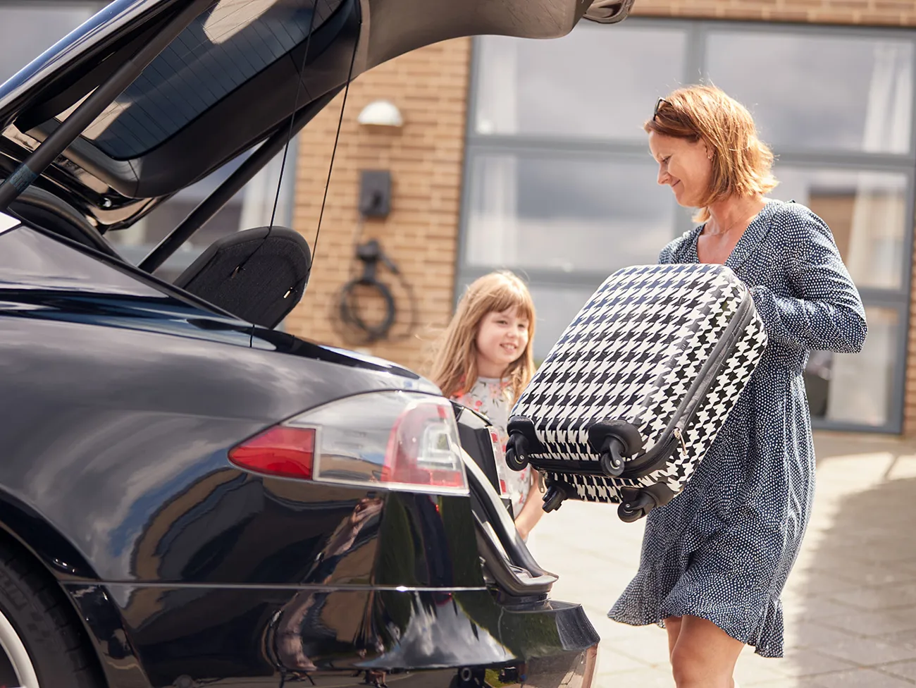 Kvinde løfter en kuffert ind i elbilens bagagerum, mens en lille pige står ved siden af bilen og kigger med