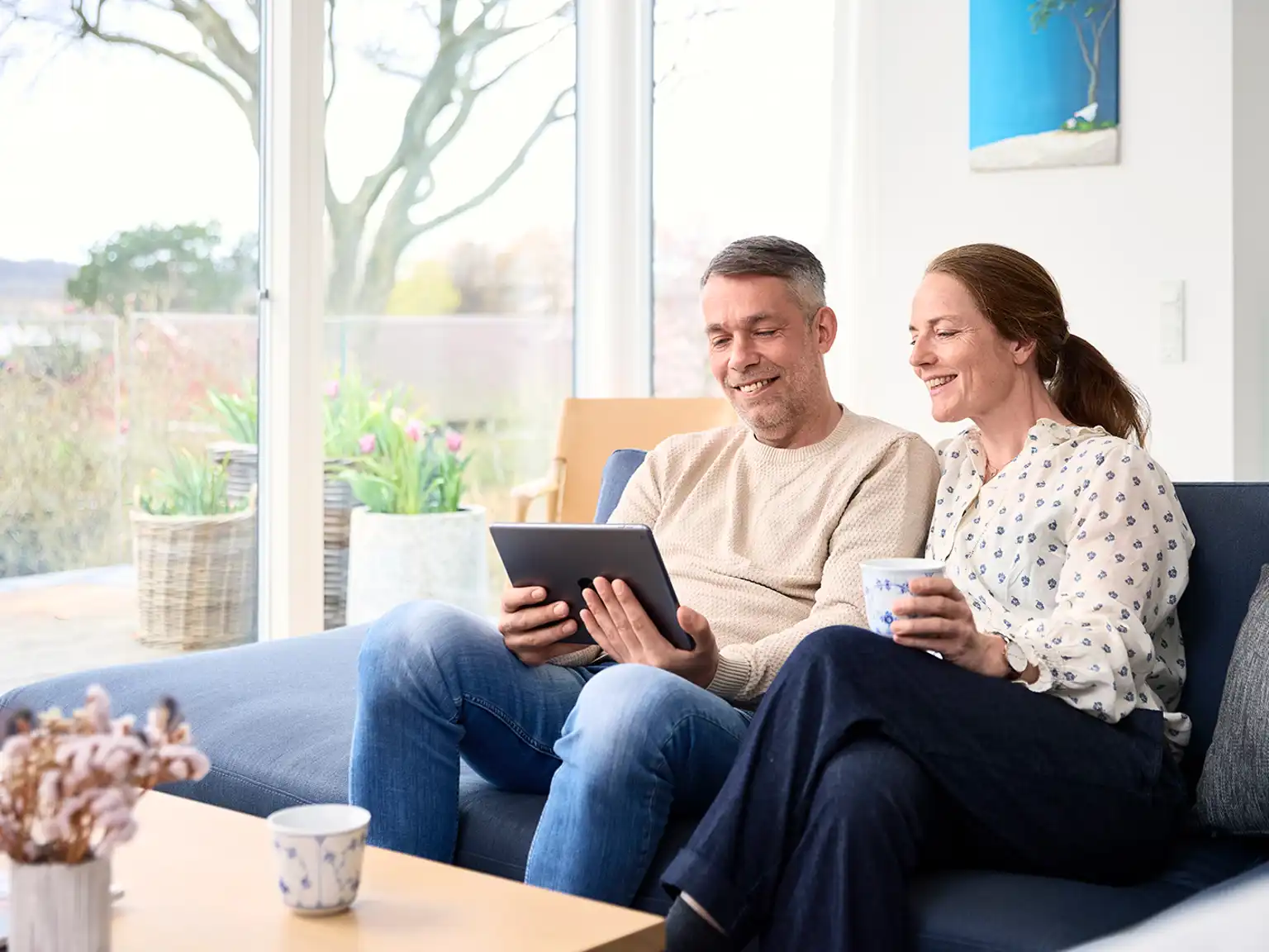 Mand og kvinde sidder sammen i sofaen og smiler manden holder en tablet som de begge kigger på og kvinden holder en kop