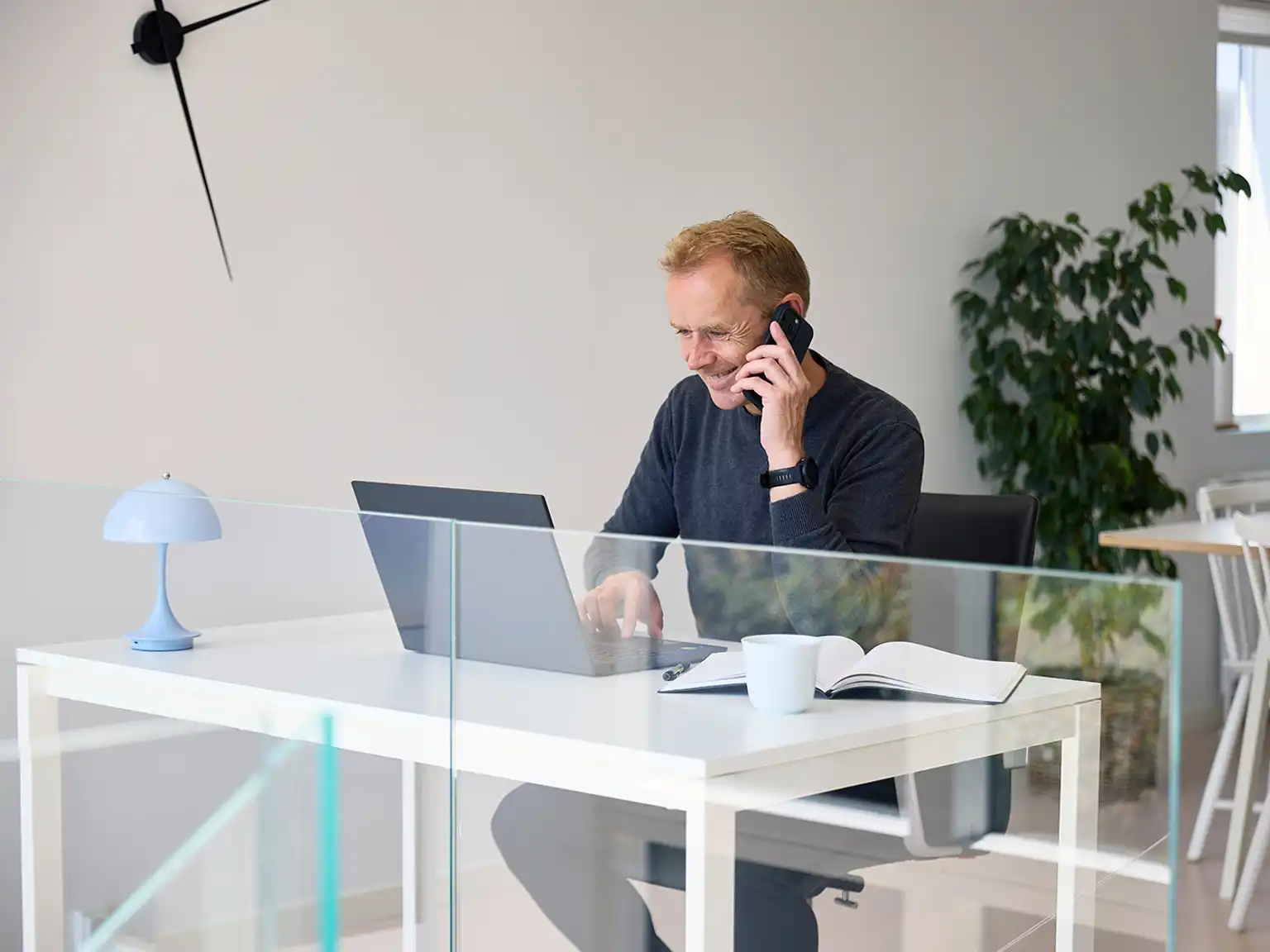 Mand sidder ved skrivebord med mobil i hånden og taster på sin computer
