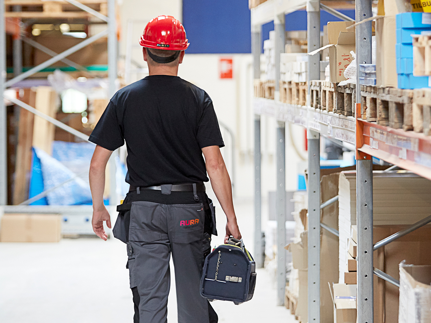Ansat ved AURA Installation går med værktøjskasse og sikkerhedshjelm gennem en lagerhal