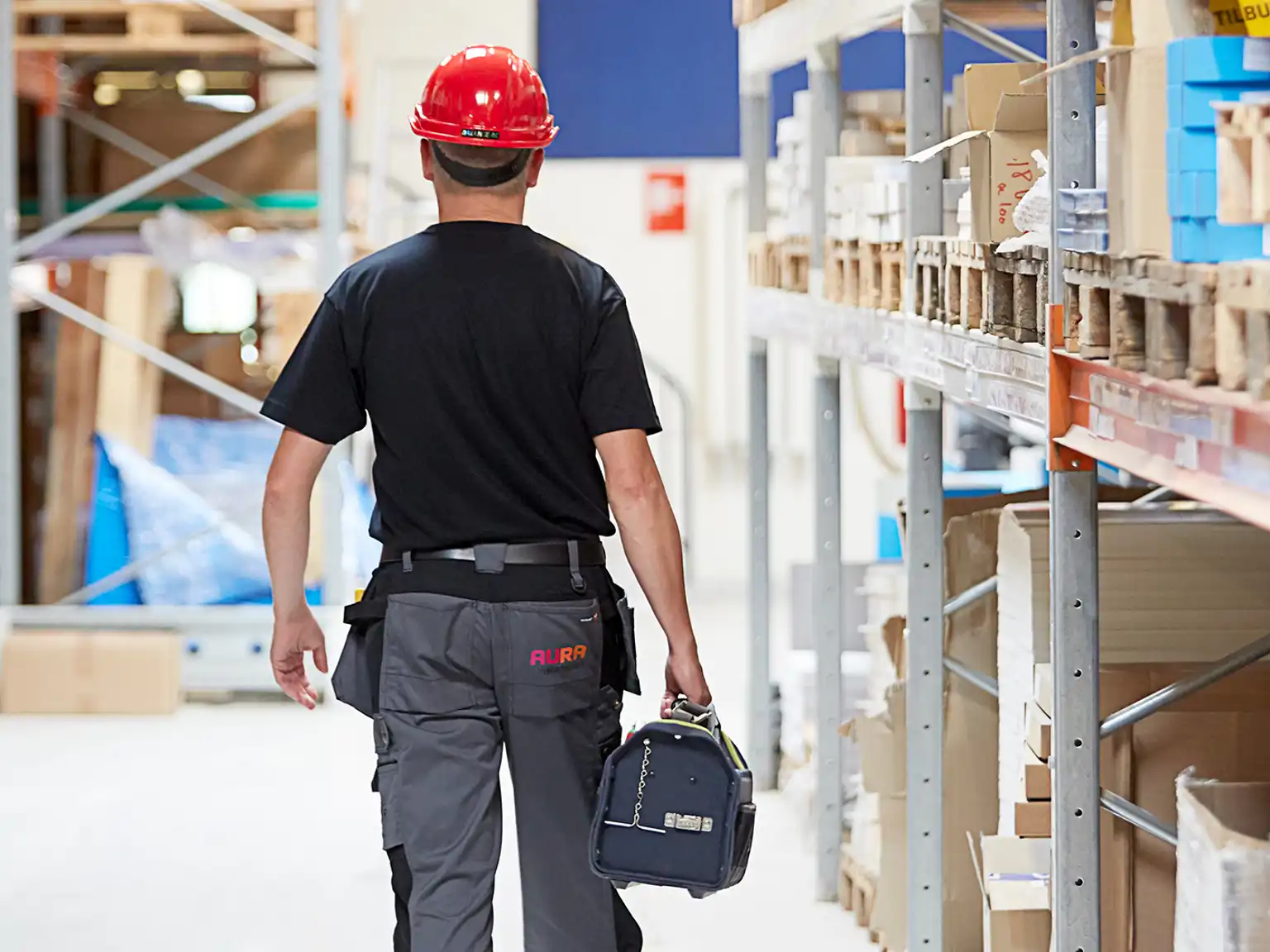 Ansat ved AURA Installation går med værktøjskasse og sikkerhedshjelm gennem en lagerhal