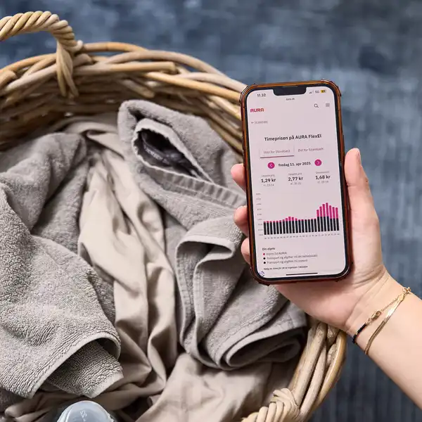 Mobil med overblik over elpriserne time for time holdes i en hånd ind over en vasketøjskurv fyldt med vasketøj