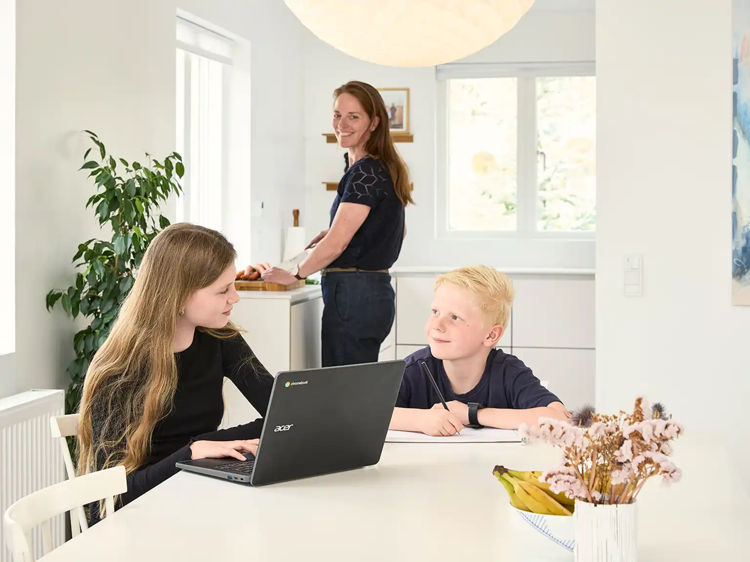 Pige og dreng sidder ved spisebordet og laver lektier mens deres mor står i køkkenet og laver mad